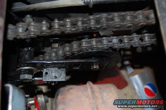Close-up of two drive chains and sprockets, one above the other. Part of a rusty exhaust pipe is visible below