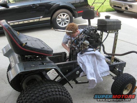 Person working on a Craftsman lawn tractor chassis with engine exposed. Black Ford Mustang in background