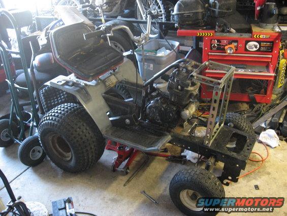 All-terrain vehicle on jack stands, undergoing repairs in a garage. Visible engine, large rear tires, and metal frame