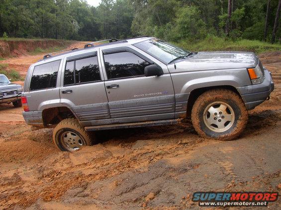 Silver Jeep Grand Cherokee stuck in mud, front wheel submerged. Another vehicle visible to the left