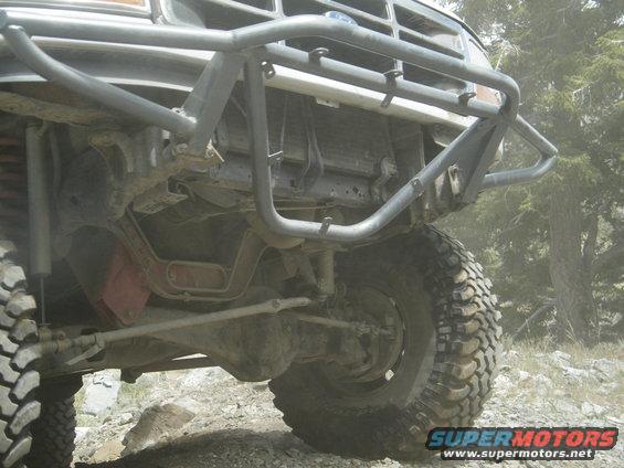 Front axle, suspension, and tire of a modified truck with a brush guard. Off-road tires and dusty conditions visible