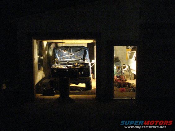 Blue truck with hood open, front view, in a garage. Another white vehicle is visible in an adjacent bay