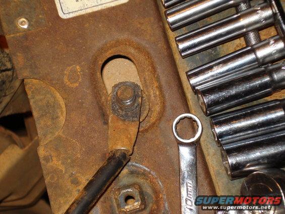 Close-up of a rusty metal bracket with a bolt and black wire attached. A 10mm wrench and socket set are visible