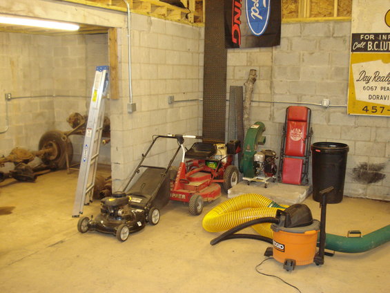 Garage interior with two lawnmowers, a leaf blower, a shop vac, a ladder, and some vehicle axles