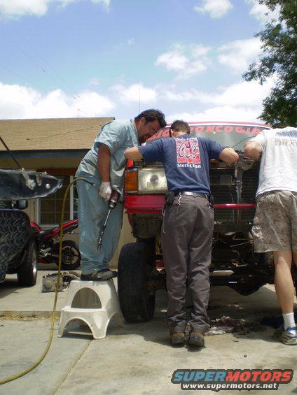 Three guys are working on the front of a red truck. One is on a step stool with a tool