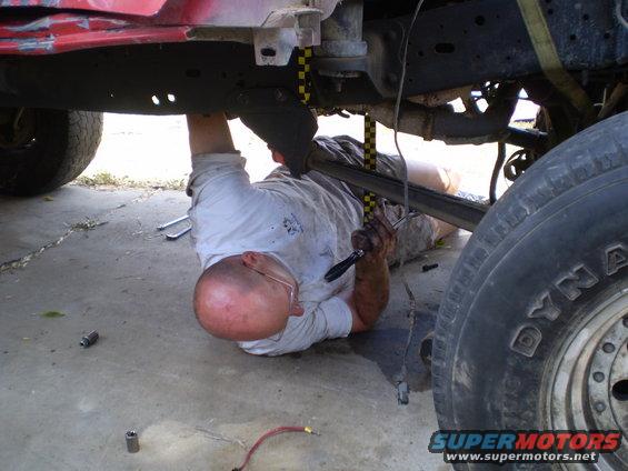 Man with glasses and bald head works on vehicle undercarriage, red paint visible on frame