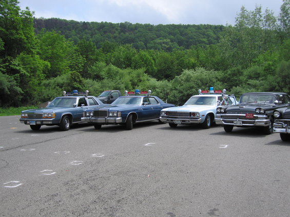 Four police cars parked in a lot, three blue and one black, with trees in the background