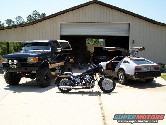 Lifted black Ford Bronco, silver DeLorean with gullwing doors open, and a black motorcycle parked in front of a garage