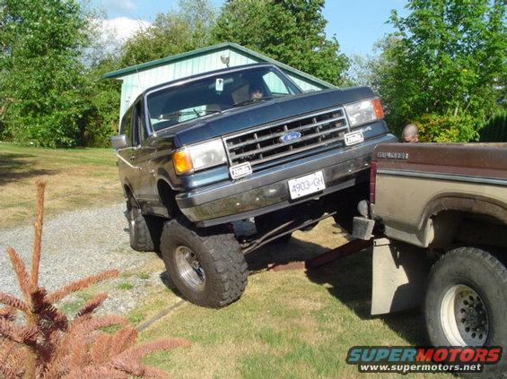 Blue Ford Bronco on a trailer hitch, next to a brown pickup truck