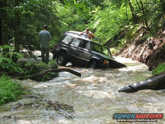 Black Jeep Cherokee XJ stuck in a stream, front end submerged, two people visible