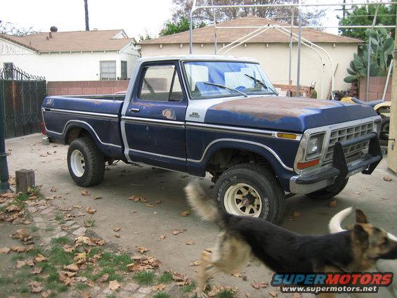 Blue and silver pickup truck with a rusty hood, parked on a dirt driveway. The truck has a chrome grille and a black brush guard