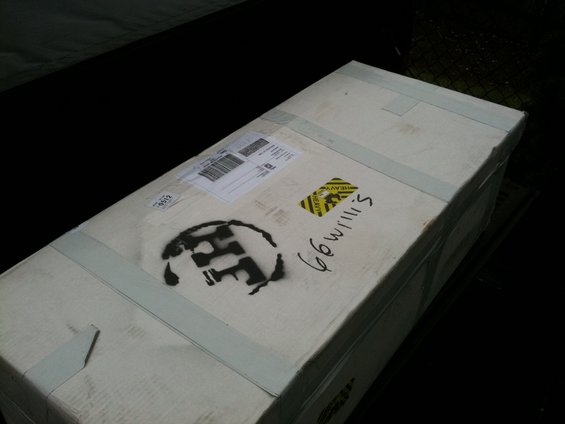 Cardboard shipping box with tape, shipping label, 'HEAVY' sticker, and a stenciled logo