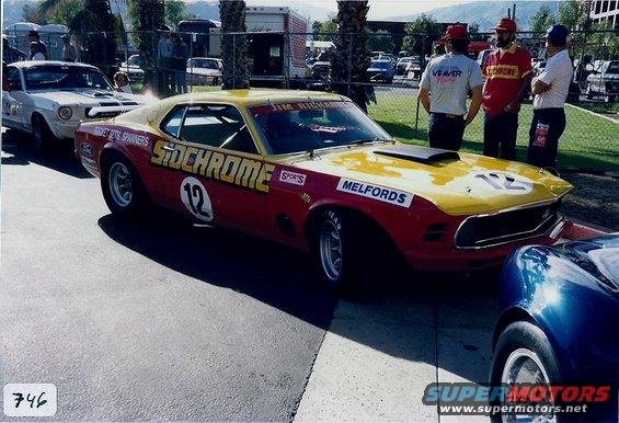 Red and yellow race car with "SIDCHROME" and "12" on the door, parked next to a white Mustang and a blue car