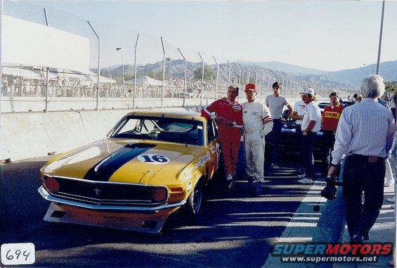 Yellow race car with black stripes and number 16 on hood, drivers in racing suits standing nearby
