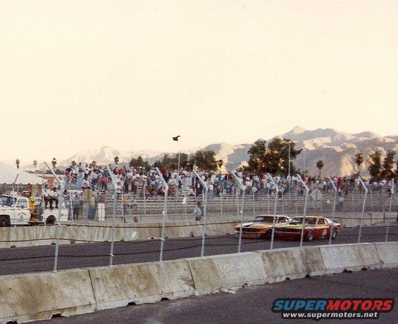 Two race cars, one orange and one red, on a track with spectators in the background