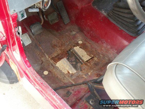 driver's side floor of a vehicle with significant rust and holes, showing pedals and part of a seat
