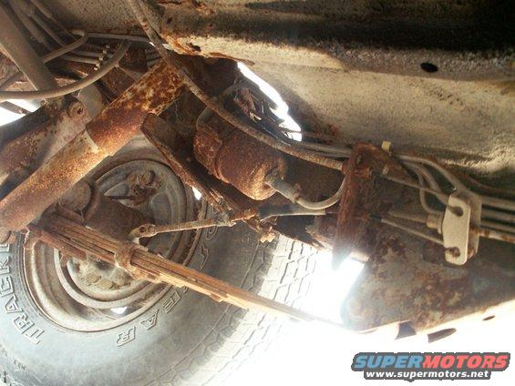 Underside of a rusty vehicle showing leaf springs, shock absorber, and fuel lines