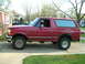 1994 Ford Bronco - A red 1994 Ford Bronco is seen from the side, showing rust damage and a dark-colored pickup truck in the background.