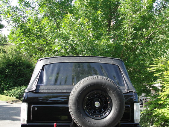 Rear view of a black SUV with a soft top and a spare tire mounted on the back. Trees are in the background
