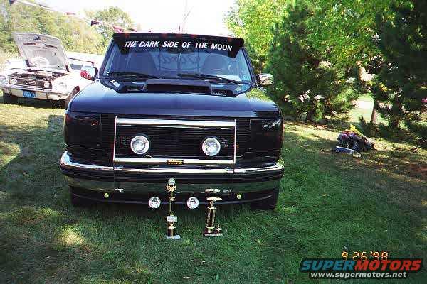 Black Ford truck with 'THE DARK SIDE OF THE MOON' banner on windshield, trophies in front. Another car with hood up in background