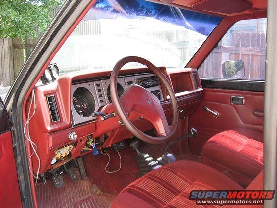 Interior of a red truck, showing the dashboard, steering wheel, and seats. Wires are visible under the dash