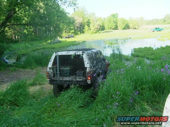 Mud-covered SUV with open rear, parked in tall grass near water; green truck in background