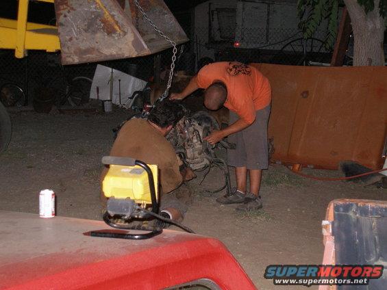 Two guys working on an engine that's being lowered by a yellow loader. A red truck is in the foreground