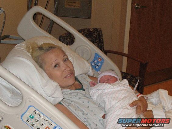 Woman in hospital gown holding a newborn baby in a hospital bed, with a "SUPERMOTORS.NET" watermark