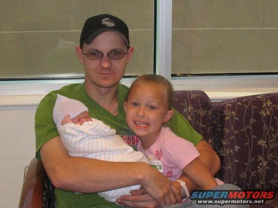 Man in green shirt and baseball cap holds a swaddled newborn; a smiling young girl in a pink shirt sits next to him