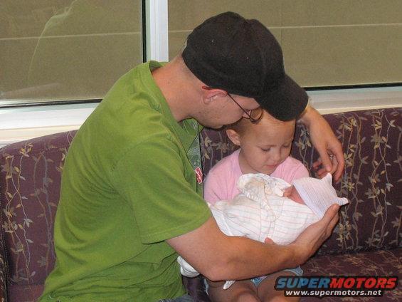 Man in green shirt and black cap holds a swaddled newborn, with a young girl looking on