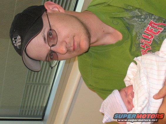 Man in a green shirt and black baseball cap holds a newborn baby wrapped in a blanket