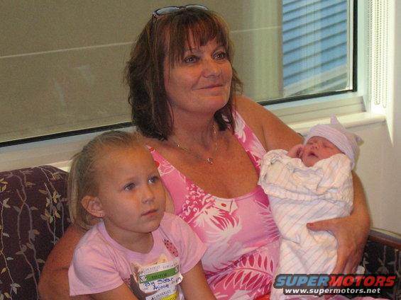 Woman in a pink floral dress holding a swaddled newborn and a young girl in a pink shirt