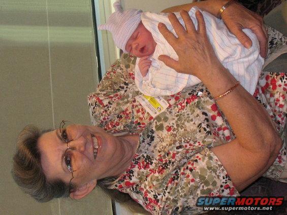 Woman wearing glasses and a floral shirt smiles while holding a swaddled newborn baby with a hat