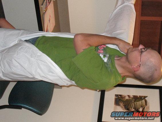 Person in green t-shirt wrapped in white sheet, lying down. Framed picture visible below