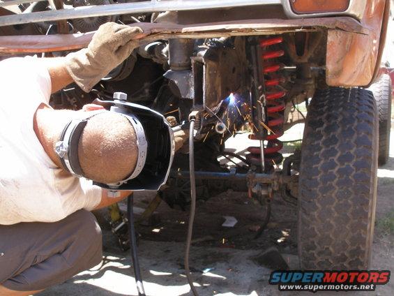 Man in welding mask works on rusty vehicle frame near red coil spring and large tire