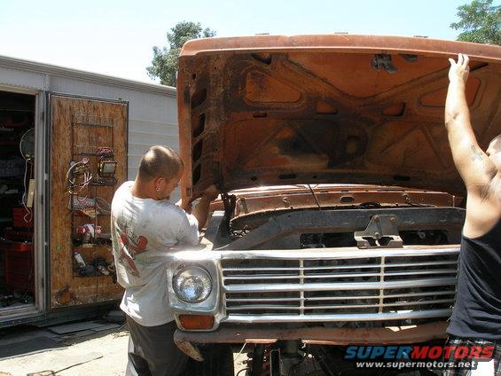 Orange truck front with hood open, men working on engine. Grille suggests early 1970s