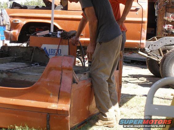 Two guys working on an orange truck bed. Another orange truck is behind them
