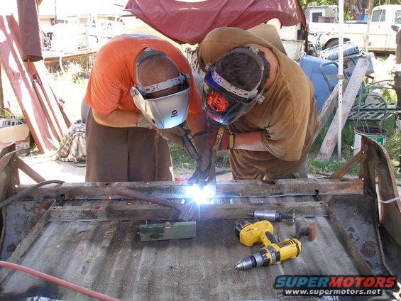 Two guys welding on what looks like a truck bed frame. A yellow drill is nearby