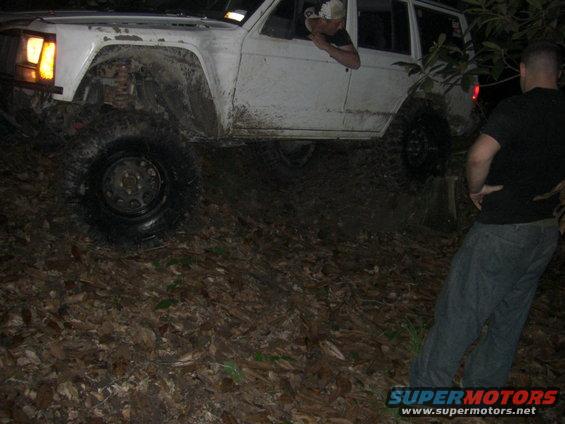 White Jeep Cherokee XJ with large tires on a muddy trail, driver visible through window