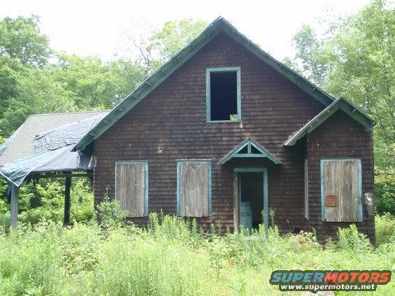 Dilapidated wooden house overgrown with weeds, boarded windows, and a tarp on the roof