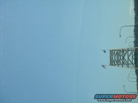 Bridge structure with two American flags and multiple streetlights against a clear blue sky. Supermotors.net logo visible