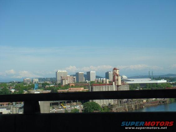 City skyline with industrial structures and a river, mountains visible on the horizon under a blue sky
