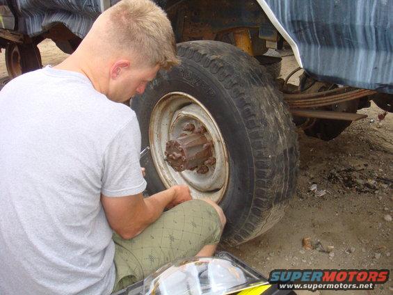 Man in grey t-shirt and green patterned shorts working on the rear axle and wheel of a vehicle