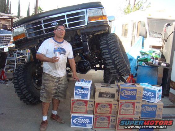 Guy holding beer next to a heavily modified Ford truck with huge tires. Another Ford truck is in the background