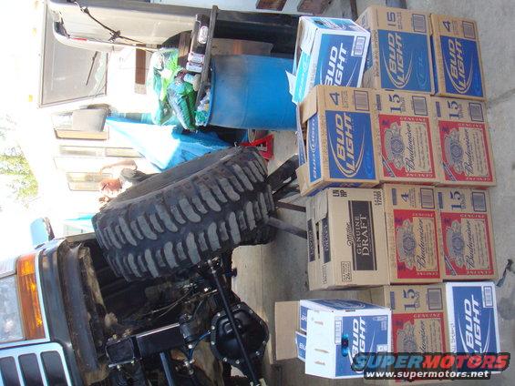 Front end of a vehicle with a large tire resting on it, surrounded by cases of beer