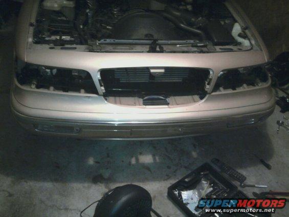 Car front end, hood open, grille visible, headlights missing. Tools and a toolbox are on the floor