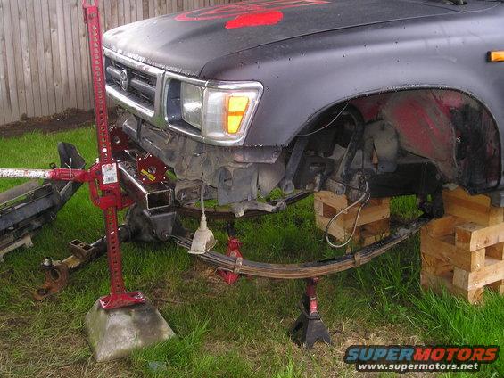 Dark Toyota truck front end lifted by a Hi-Lift jack and jack stands, showing leaf spring suspension