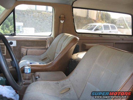 Tan interior of a 1980s SUV with bucket seats, steering wheel, and a water bottle. Another white SUV is visible through the rear window