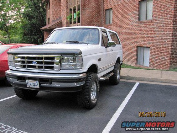 White Ford Bronco parked in a lot, front 3/4 view. Chrome grille, aftermarket wheels, and a bug deflector are visible