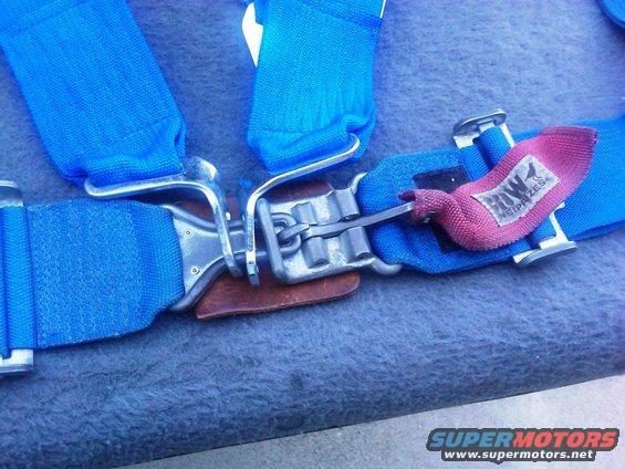 Blue racing harness buckle with pink tag reading 'ROW NEIPRIZES'